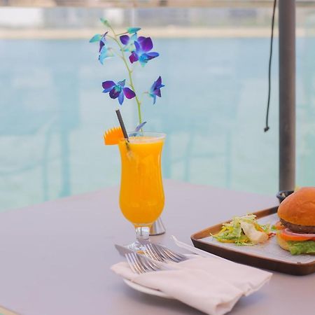 Grand Swiss-Belhotel Waterfront Seef Sanabis Exterior photo The photo features a refreshing scene with a vibrant orange drink garnished with a slice of orange and purple flowers, served in a glass. Next to it, there is a tray containing a hamburger with lettuce and possibly other condiments, accompanied by a 