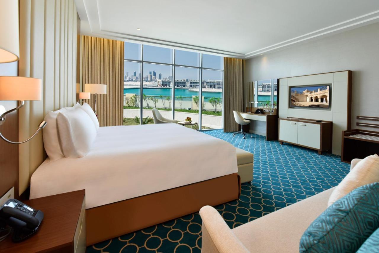 Grand Swiss-Belhotel Waterfront Seef Sanabis Exterior photo The photo shows a luxurious hotel room. It features a large bed with white linens, and there is a comfortable armchair and a small sofa in the corner. The room has large windows that offer a view of the outdoors, likely facing a body of water. The ca
