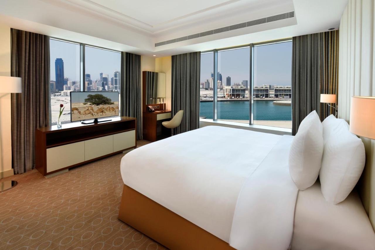 Grand Swiss-Belhotel Waterfront Seef Sanabis Exterior photo The photo shows a luxurious hotel room featuring a large bed with white pillows and a neatly made bedspread. The room has floor-to-ceiling windows that offer a view of a city skyline, suggesting a vibrant urban setting. There is a modern television m