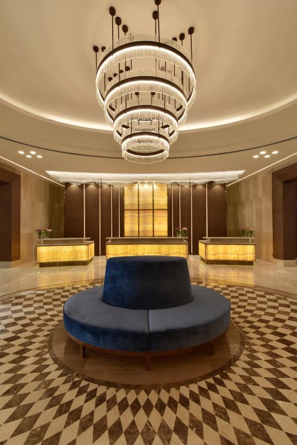 Grand Swiss-Belhotel Waterfront Seef Sanabis Exterior photo The photo shows an elegant hotel lobby. It features a circular seating area in the center with a plush, dark blue upholstered bench. Surrounding this seating area is a patterned floor made of light and dark tiles. Behind the seating area, there are t