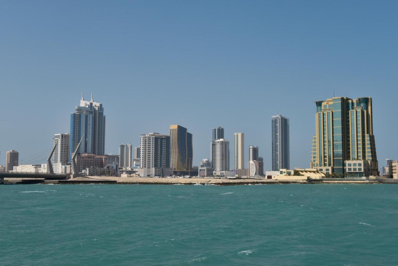 Grand Swiss-Belhotel Waterfront Seef Sanabis Exterior photo The photo shows a skyline consisting of several tall buildings and skyscrapers along a waterfront. The buildings vary in design and height, showcasing modern architecture typical of urban areas. The water in the foreground is a vibrant turquoise colo