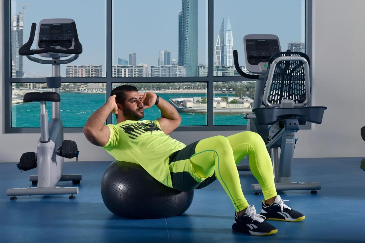 Grand Swiss-Belhotel Waterfront Seef Sanabis Exterior photo The photo shows a man exercising in a gym setting. He is performing a sit-up while balancing on an exercise ball. He is wearing a bright neon yellow athletic outfit and black shoes. In the background, large windows provide a view of a city skyline an