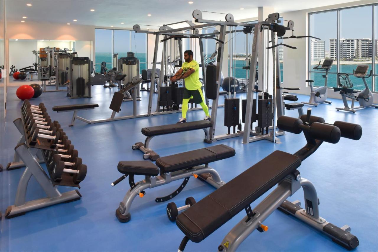 Grand Swiss-Belhotel Waterfront Seef Sanabis Exterior photo The photo shows a modern gym interior with a variety of fitness equipment. In the foreground, there is a weight bench and a set of dumbbells arranged neatly. To one side, there is a multi-station workout machine where a person is performing an exerci