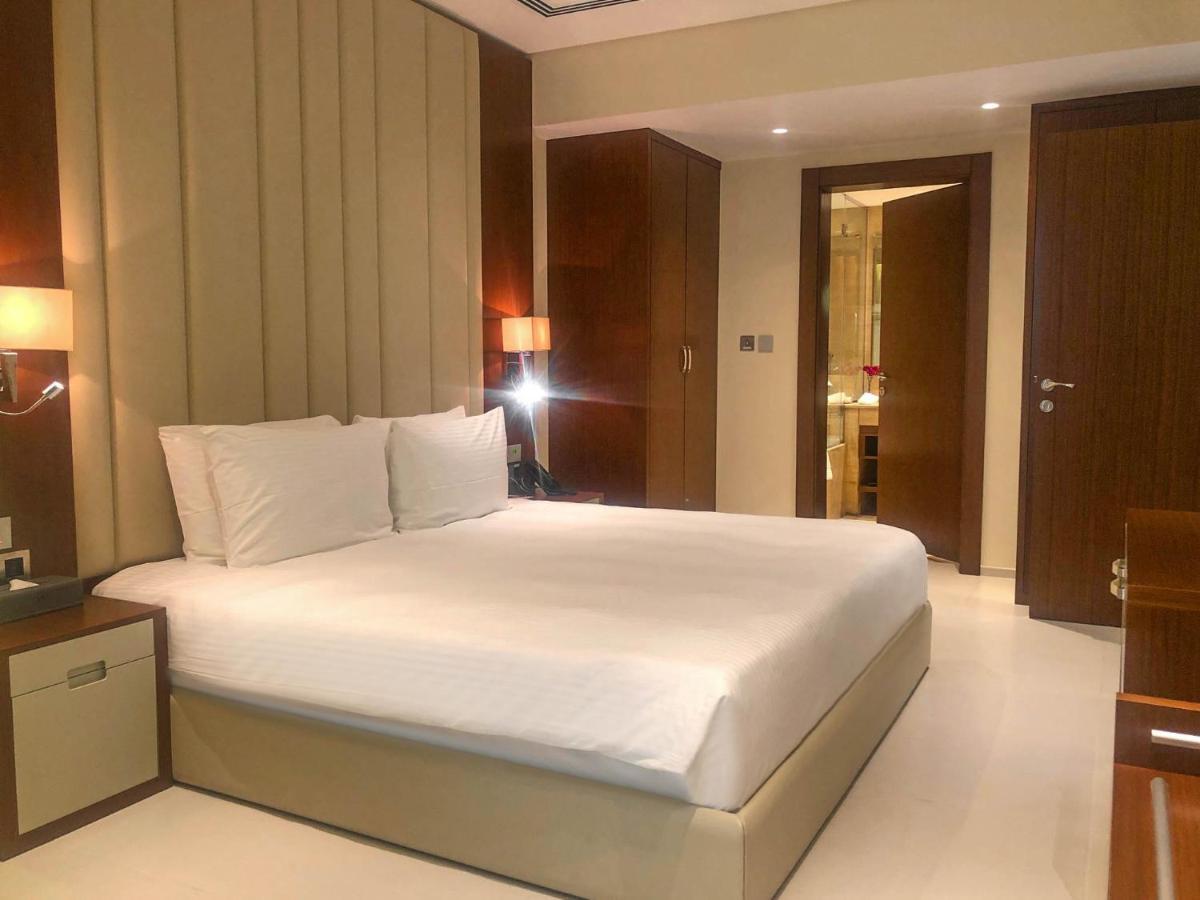 Grand Swiss-Belhotel Waterfront Seef Sanabis Exterior photo The photo shows a modern hotel room. It features a large bed with white linens and two pillows, positioned against a wall with vertical beige panels. There are nightstands on either side of the bed, and each has a lamp. The room has a clean and minim