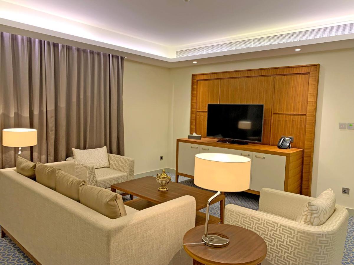 Grand Swiss-Belhotel Waterfront Seef Sanabis Exterior photo The photo shows a modern living room area. There are two sofas arranged around a coffee table, which has a decorative item on it. A lamp is positioned next to one of the sofas, providing ambient lighting. In the background, there is a large flat-scre