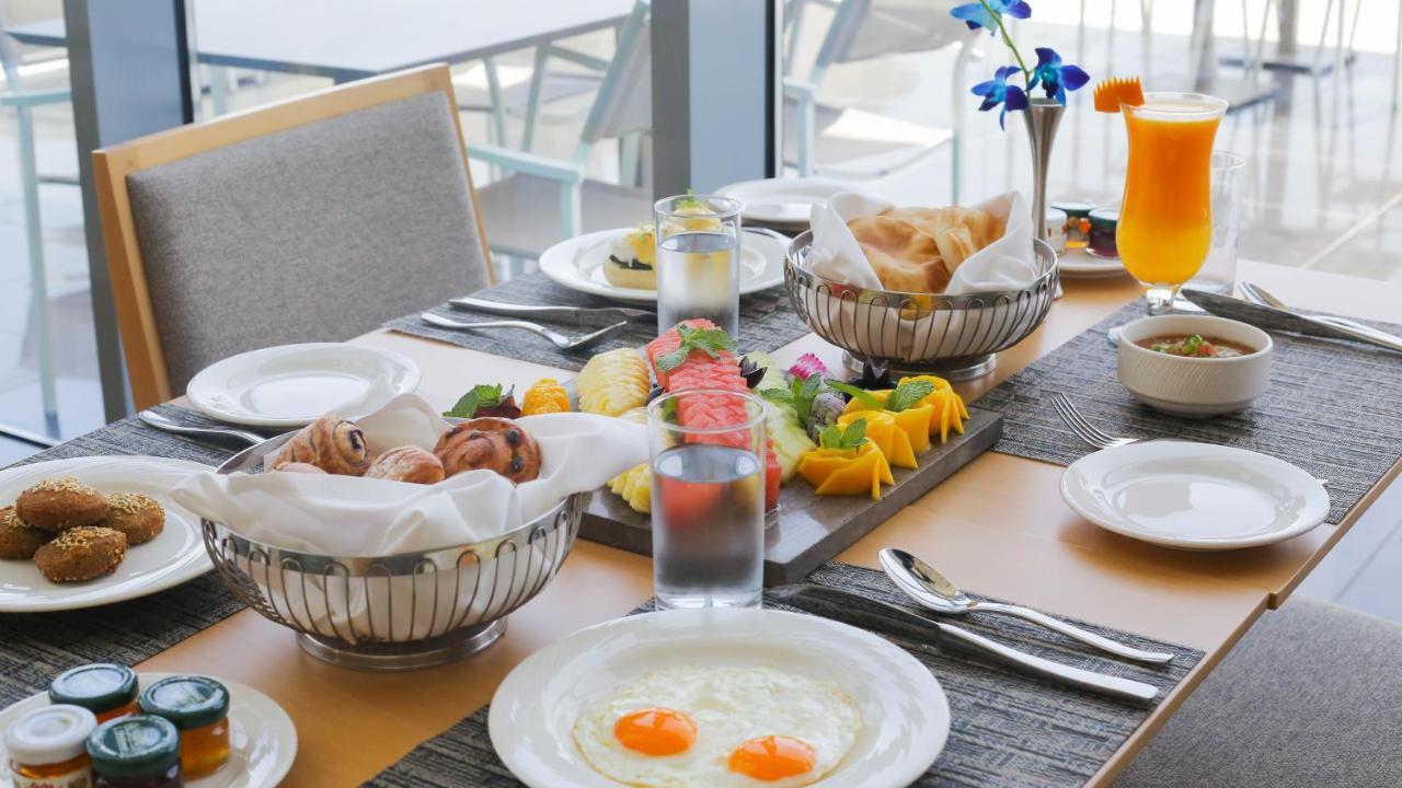 Grand Swiss-Belhotel Waterfront Seef Sanabis Exterior photo The photo displays a beautifully arranged breakfast table. At the center, there's an assortment of fresh fruits, including watermelon and mango, presented on a wooden platter. Around the table, there are dishes containing pastries and bread, neatly p