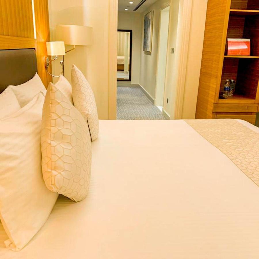 Grand Swiss-Belhotel Waterfront Seef Sanabis Exterior photo The photo shows a neatly made bed with a light-colored blanket and decorative pillows. In the background, there is a hallway leading to other room areas. The walls are painted in neutral tones, and there are elements of modern decor, such as a bedsid