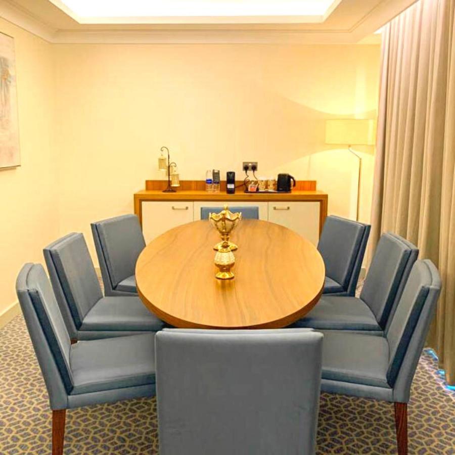 Grand Swiss-Belhotel Waterfront Seef Sanabis Exterior photo The photo shows a modern dining or conference room setup. It features a round wooden table surrounded by several blue upholstered chairs. In the center of the table, there is a decorative golden piece, likely a vase or trophy. To one side of the room